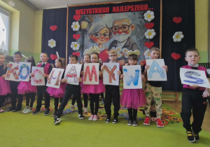Dzieci recytują wiersze trzymając przed sobą karty z napisem KOCHAMY WAS