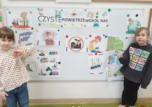 Dwie dziewczynki stoją przy tablicy z ilustracjami przedstawiającymi zanieczyszczenia powietrza i sposoby jak je ograniczać
