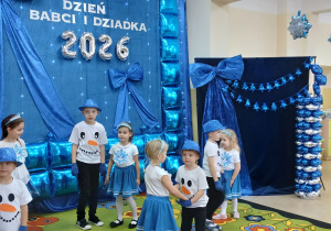 Dzieci w biało niebieskich strojach na tle dekoracji prezentują taniec.