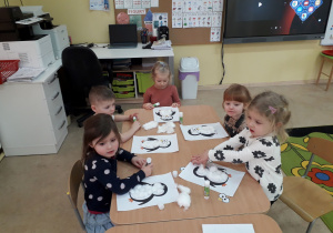 Grupa młodszych dzieci przy stoliku ozdabia watą sylwetę pingwina.