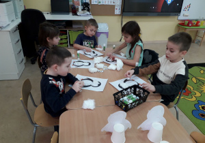 Grupa starszaków przy stoliku ozdabia watą sylwetę pingwina.