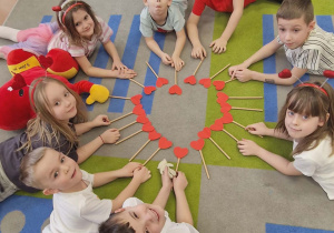 Dzieci leżą na dywanie, w środku serce z ułożonych serc na patyku