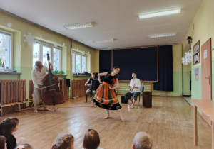 Panowie grają na instumentach a pani ubrana w łowicki strój prezentuje taniec.