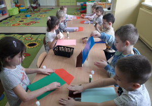 Dzieci przy stolikach projektują i sklejają domki z figur geometycznych.