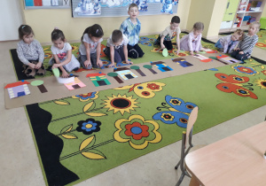 Dzieci na dywanie prezentują makietę wioski geometrycznej wykonanej z figur geometrycznych.