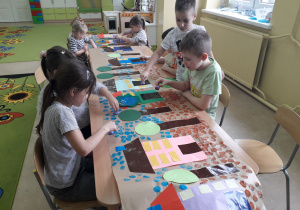 Dzieci przy stolikach stemplują okrągłymi pomponami makietę geometrycznej wioski.