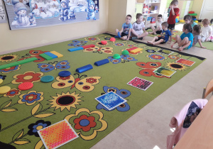 Na dywanie rozłożona ścieżka sensoryczna. Dzieci przygotowują się do jej pokonania.