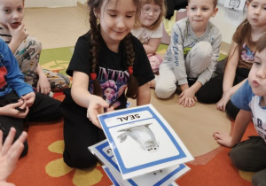 Dzieci siedzą w kole, w środku dziewcznka układa więżę z obrazków i kubeczków podczas zabawy w języku angielskim.