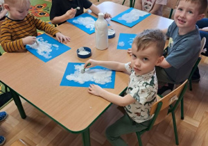Dzieci siedzą przy stoliku i wykonują pracę plastyczną z użyciem farby.