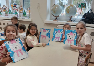 Dziewczynki siedzą przy stoliku i prezentują wykonaną pracę plastyczną przestawiającą niedźwiedzia polarnego.