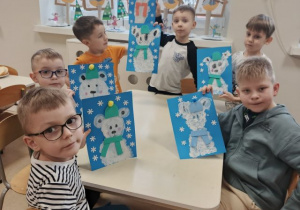 Chłopcy siedzą przy stoliku i prezentują wykonaną pracę plastyczną przestawiającą niedźwiedzia polarnego.