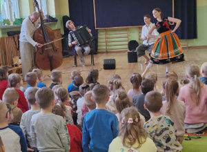 Dzieci siedzą na dywanie. Przed nimi muzycy grają na instrumentach i pani ubrana w łowicki strój tańczy