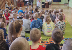 Dzieci siedzą na dywanie. Przed nimi muzycy grają na instrumentach i pani ubrana w białą sukienkę trzyma koszyk z ziołami