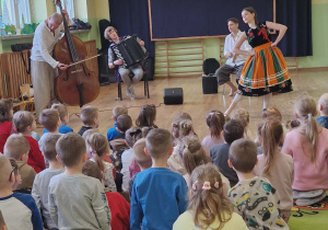 Dzieci siedzą na dywanie. Przed nimi muzycy grają na instrumentach i pani ubrana w łowicki strój tańczy