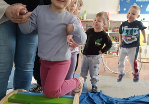 Dziecko przechodzi przez drabinkę z pomocą nauczyciela, w tle dzieci ustawione w rzędzie