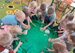 Dzieci siedzą w kole i próbują wydobyć dinozaury z zamrożonej bryły lodu w kształcie jaja.