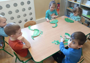 Dzieci siedzą przy stoliku i tworzą dinozaury z papierowych talerzyków pomalowanych na zielono.