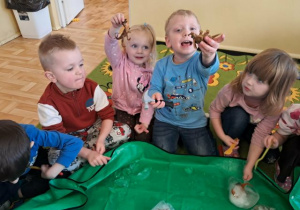 Dzieci siedzą w kole i próbują wydobyć dinozaury z zamrożonej bryły lodu w kształcie jaja.