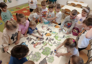 Przedszkolaki stoją dookoła stołu i przyklejają obrazki dinozaurów na dużym brystolu.