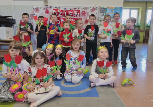 Dzieci pozuje do zdjęcia grupowego na tle dekoracji z kwiatami zrobionymi dla mam i sióstr