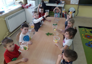 Dzieci przy stolikach prezentują wykonany napój z wody, owoców i świeżych ziół.