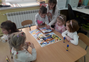 Dziewczynki przy stoliku składają w całość plakat "W marcu jak w garncu".