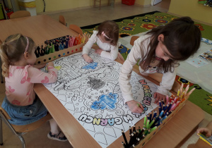 Dziewczynki przy stoliku kolorują duży plakat "W marcu jak w garncu".