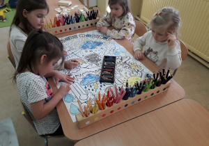 Dziewczynki przy stoliku kolorują duży plakat "W marcu jak w garncu".