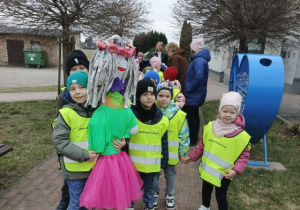 Przedszkolaki ubrane w kamizelki odblaskowe, ustawione parami przygotowują się do spaceru. Chłopiec trzyma marzannę.