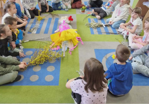 Dzieci siedzą na dywanie w kole. Przed nimi przyniesione z domu wiosenne rośliny i bazie.