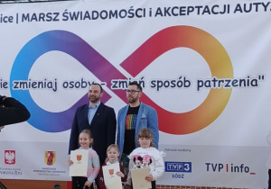 Dzieci przedszkolne z wręczonymi dyplomami i nagrodami stoją z przedstawicielami wydarzenia na tle dekoracji.