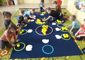 Dzieci na dywanie podczas zabawy matematycznej dopasowują ilość piłeczek do cyfry napisanej na sylwecie kurki.