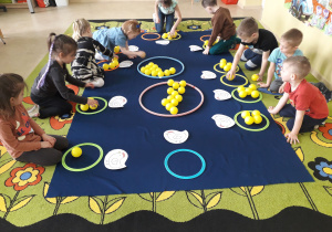 Dzieci na dywanie podczas zabawy matematycznej dopasowują ilość piłeczek do cyfry napisanej na sylwecie kurki.