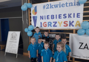Dzieci w niebieskich koszulkach pozują do zdjęcia na tle baneru Niebieskie Igrzyska.