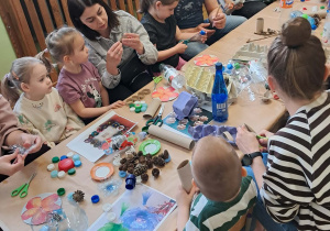 Dzieci z rodzicami siedzą przy stołach i wykonują kwiaty z materiałów ekologicznych.