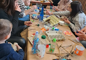 Dzieci z rodzicami siedzą przy stołach i wykonują kwiaty z materiałów ekologicznych.
