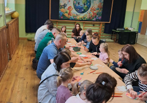 Dzieci z rodzicami siedzą przy stołach i wykonują podstawki z materiałów ekologicznych.