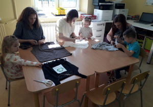 Warsztaty ekologiczne. Mamy z dziećmi przy stolikach podczas wykonywania torby ekologicznej na zakupy z niepotrzebnej koszulki.