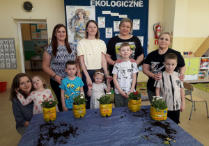 Mamy i dzieci pozują do zdjęcia, prezentując kwiaty w ekologicznych donicach.