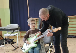 Chłopiec siedzi na krześle i gra na gitarze elektrycznej przy pomocy muzyka.