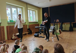Dzieci siedzą na dywanie i słuchają utworu granego przez muzyka na ukulele.
