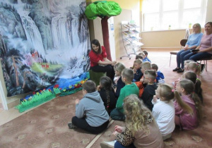Dzieci siedzą na dywanie w bibliotece i słuchają bajki czytanej przez panią.