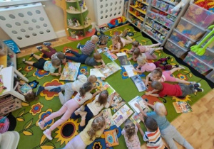 Przedszkolaki leżą na brzuszkach na dywanie i oglądają książeczki.