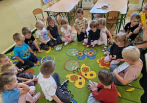 Przedszkolaki siedzą w kole na dywanie i ozdabiają kropeczkami małe obręcze.