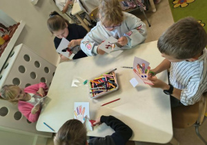 Dzieci siedzą przy stoliku i wycinają kolorowe dłonie z papieru.