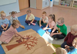 Dzieci siedzą w kole. W środku praca plastyczna z drzewem życzliwości. Dziecko przykleja listek.
